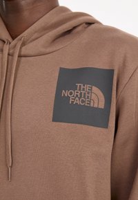 Detailní záběr na hnědou mikinu s kapucí a stahovacími šňůrkami, vyznačující se černým čtvercovým nášivkem s textem a emblémem "The North Face" na hrudi.