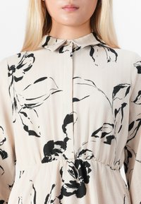Femme blonde portant une robe beige avec des motifs floraux noirs, col boutonné et taille froncée, vue de la nuque à la taille.