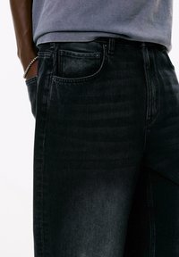 Den nederste del af torsoen har sorte slidte jeans og en grå skjorte, med den ene hånd i lommen og et sølvarmbånd på håndleddet.