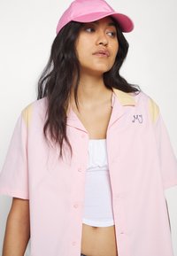 Chemise rose à manches courtes avec un col crème et des initiales brodées. Associée à une casquette rose et un crop top blanc en dessous.