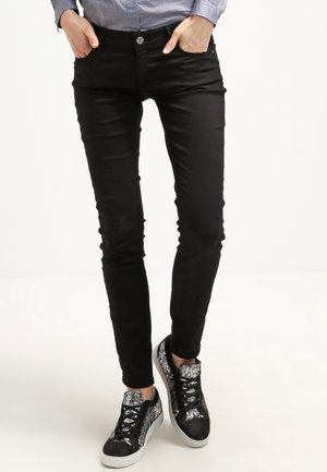 Person iført slanke sorte bukser med hænderne i lommerne og sort-hvide sneakers med mønster, stående på en lys baggrund.