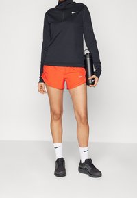 černý dlouhý rukáv s čtvrtinovým zipem, oranžové šortky, bílé logo Nike na obou, v kombinaci s černými teniskami a bílými ponožkami, drží černou láhev na vodu.