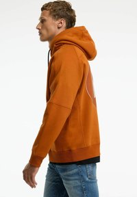 Oranje hoodie met een grote grafische print op de achterkant, voorzien van een capuchon met trekkoord en geribde manchetten. G gedragen met blauwe jeans.