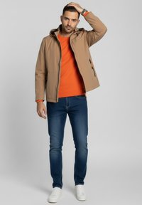 Chaqueta beige con capucha, cremallera negra y detalles naranjas sobre un suéter naranja; jeans azules y zapatillas blancas completan el conjunto.