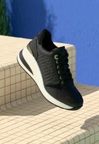 Black sneaker with white sole and green eyelets resting on beige tiled steps against a blue wall background.