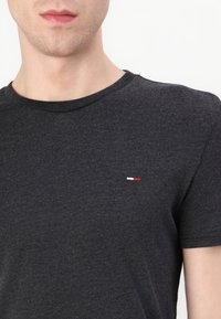 Nahaufnahme einer Person, die ein dunkelgraues T-Shirt mit Rundhalsausschnitt und einem kleinen rot-weißen Logo auf der linken Brust trägt.