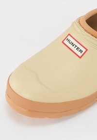 Beige színű, gumiból készült slip-on cipő narancssárga talppal, amely texturált felülettel rendelkezik, és a felső részén egy piros kerettel ellátott HUNTER logó címke található.