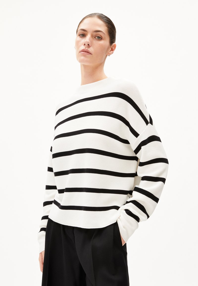 Weißer Pullover mit schwarzen horizontalen Streifen, geripptem Muster, Rundhalsausschnitt und überschnittenen Schultern, kombiniert mit schwarzen Hosen.