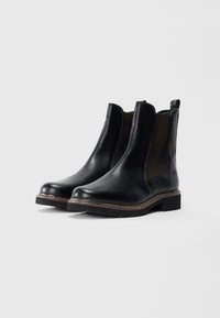 Bottines Chelsea en cuir noir avec panneaux élastiques marron sur les côtés, languettes de tirage et semelles en caoutchouc épaisses sur fond blanc.