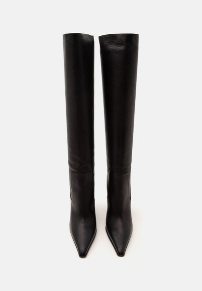 Paire de hautes bottes en cuir noir avec bouts pointus, debout sur un fond blanc, vue de face.