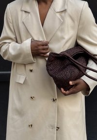 Beige double-breasted coat with a notched collar and gold buttons, held with a hand grasping a dark woven handbag with two straps.