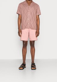 Man i rosa mönstrad kortärmad skjorta, enfärgade ljusrosa shorts och svarta lädersandaler på plain bakgrund.