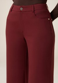 Bordeaux wijd-uitlopende broek gemaakt van een gladde stof, met een sluiting met een voorknop, traditionele zakken en subtiele accenten van zilverkleurig beslag.