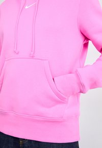 Roze hoodie gemaakt van zachte stof, met een voorkant zak, een capuchon met trekkoord, geribde manchetten en zoom, en een klein wit logo op de borst.