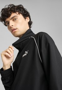 Black track jacket made of smooth fabric, features a high collar, zip closure, white accents, and Puma logo on the chest.