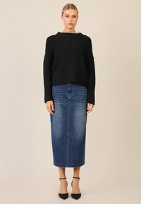 Schwarzer gerippter Strickpullover mit weiten Ärmeln; kombiniert mit einem Denim-Maxikleid mit Nietenverzierungen und gerader Silhouette. Schwarze Pumps.