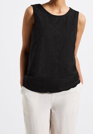 Donna con indosso un top nero ricamato senza maniche con orlo smerlato e pantaloni larghi di colore chiaro con le mani nelle tasche.