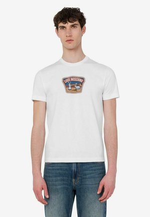 Młody mężczyzna z krótkimi kręconymi włosami, ubrany w białą koszulkę Love Moschino z kolorową grafiką oraz niebieskie jeansy, stojący na jednolitym tle.