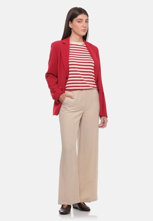 Donna in piedi che indossa un blazer rosso, una camicia a righe rosse e bianche, pantaloni beige a gamba larga e scarpe nere, con una mano in tasca.