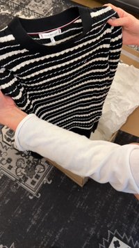 Des mains tiennent un pull Tommy Hilfiger à rayures noires et blanches au-dessus d'une boîte en carton ouverte sur un sol à motifs.
