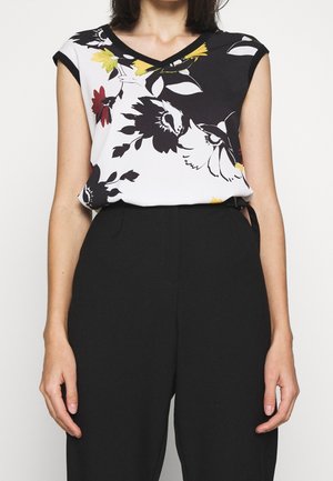 Femme portant un pantalon noir sans manches et un haut blanc sans manches imprimé de motifs floraux et d'oiseaux en noir, jaune et rouge.