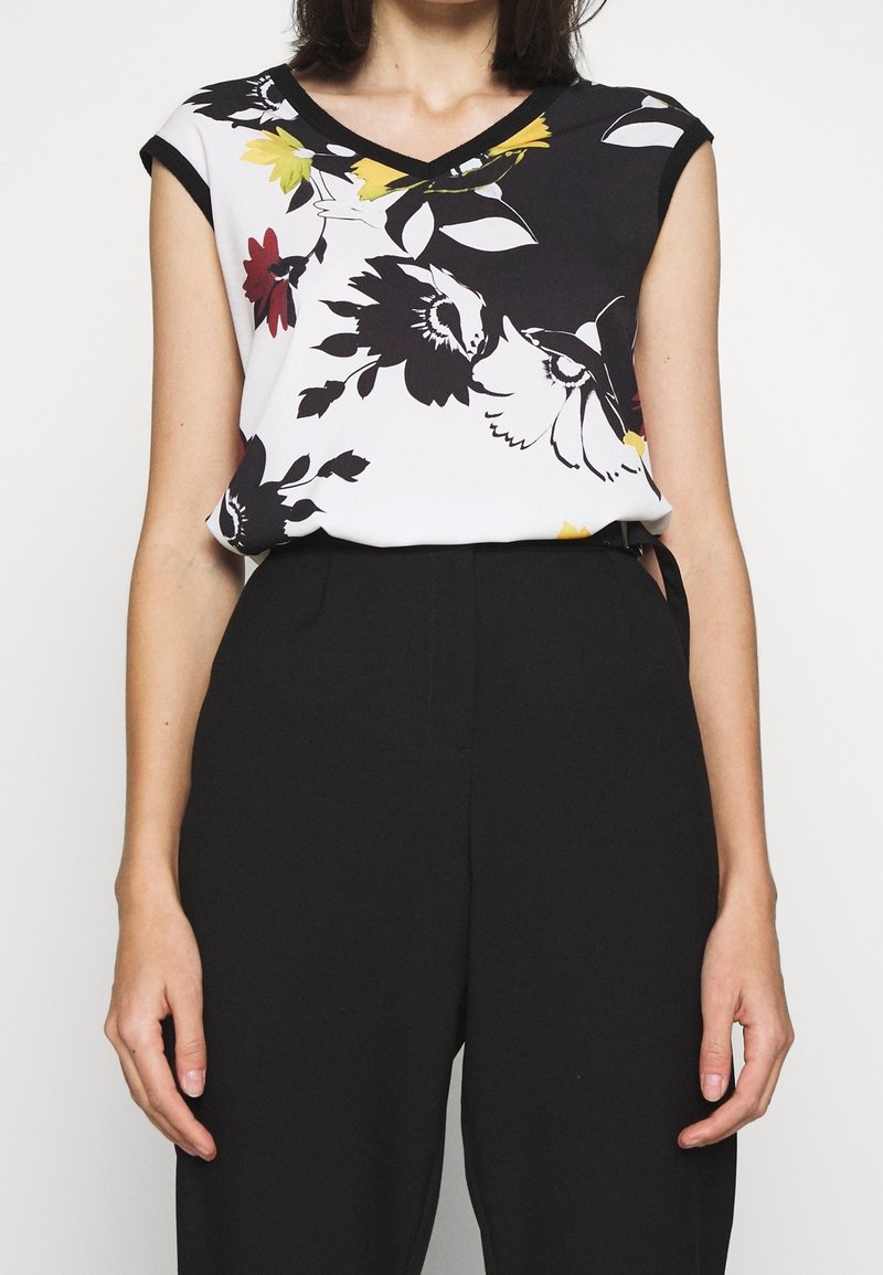 Femme portant un pantalon noir sans manches et un haut blanc sans manches imprimé de motifs floraux et d'oiseaux en noir, jaune et rouge.