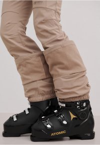 Sorte skistøvler med metallisk hardware og justerbare klips, kombinert med beige snøbukser med myk, strukturert mansjett og slankt design.