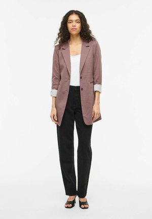 Femme portant un blazer mauve avec des manches retroussées, un haut blanc, un pantalon noir taille haute et des escarpins noirs à bouts ouverts, debout devant un fond blanc.