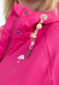 Imperméable rose vif avec fermeture éclair, boutons-pression noirs, logo parapluie blanc et cordon tressé multicolore avec perle en bois près du col.