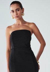 Nainen, jolla on takertunut hiustyyli, pukeutuneena olkaimettomaan mustaan mekkoon, suurikokoisiin kultaketjuihin ja kultakintaan, vaaleanharmaata taustaa vasten.