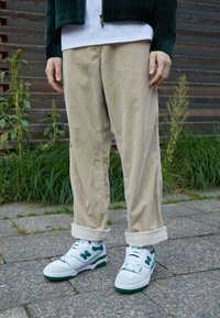 Pantaloni in velluto a coste beige con orli arrotolati, abbinati a sneaker bianche con accenti verdi e una superficie testurizzata. Look casual da strada.