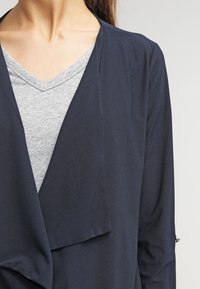 Marinblå, lättvikts cardigan med öppen framside och draperad design; lagd över en grå t-shirt med v-ringning; slät textur.