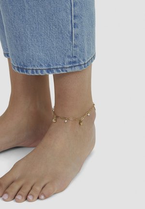 Bare feet with light pink toenails wearing a gold chain anklet with small heart and pearl charms, paired with cropped blue denim jeans.