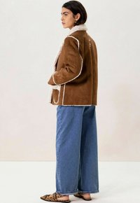Veste en daim synthétique marron avec doublure et bordure en shearling blanc, coupe détendue, portée sur un jean bleu ample et des ballerines imprimé animal.