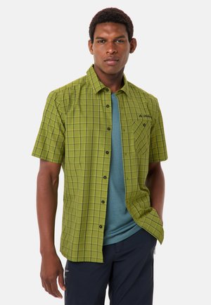 Uomo che indossa una camicia a maniche corte verde a quadri sopra una T-shirt blu e pantaloni scuri, in piedi con una mano in tasca.