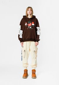 Joven de pie con una sudadera marrón con estampados de dibujos animados, pantalones cargo crema con bolsillos y botas de cordones color tostado sobre un fondo blanco.