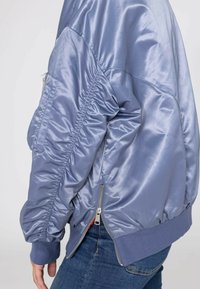 Jasnoniebieska satynowa kurtka bomberka z prążkowanymi mankietami i pasem, z bocznymi kieszeniami i zamkiem błyskawicznym na dole.