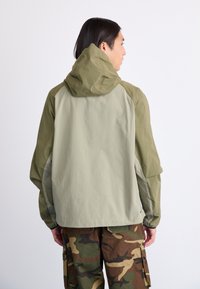 Letvægts hættejakke i to nuancer af lys olivengrøn og mørkegrøn. Har raglanærmer, glat tekstur og et minimalistisk design.