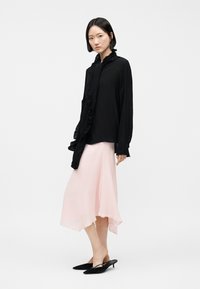 Mujer con cabello corto y negro, que lleva una blusa negra de manga larga con volantes, una falda midi color rosa pálido y zapatos negros de tacón bajo.