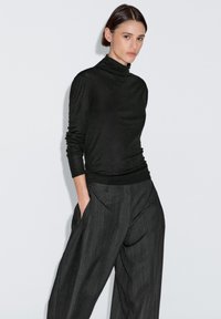 Maglione a collo alto nero in materiale leggero, abbinato a pantaloni a gamba larga grigio scuro a righe, dotati di tasche laterali e vestibilità rilassata.