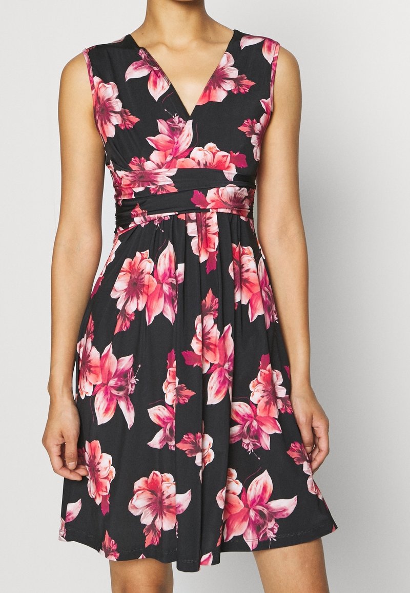 Robe noire sans manches avec imprimé floral rose et rouge, taille ajustée et col en V, portée par une personne sur un fond uni.