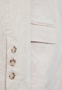 Blazer de lino beige claro con cuatro botones forrados de tela en la manga, un bolsillo y una tela texturizada y transpirable.