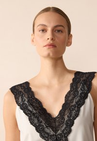 Top blanco con un escote en V profundo, que presenta un amplio borde de encaje floral negro a lo largo del escote. Textura de tela suave, diseño sin mangas.