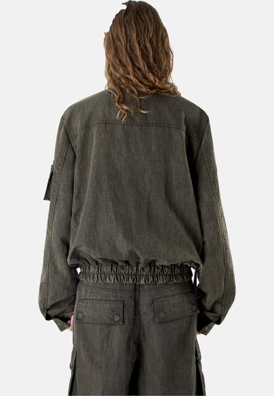 Persona con cabello largo vista desde atrás, vistiendo una chaqueta holgada de color verde oliva oscuro con cintura fruncida y pantalones cargo a juego.