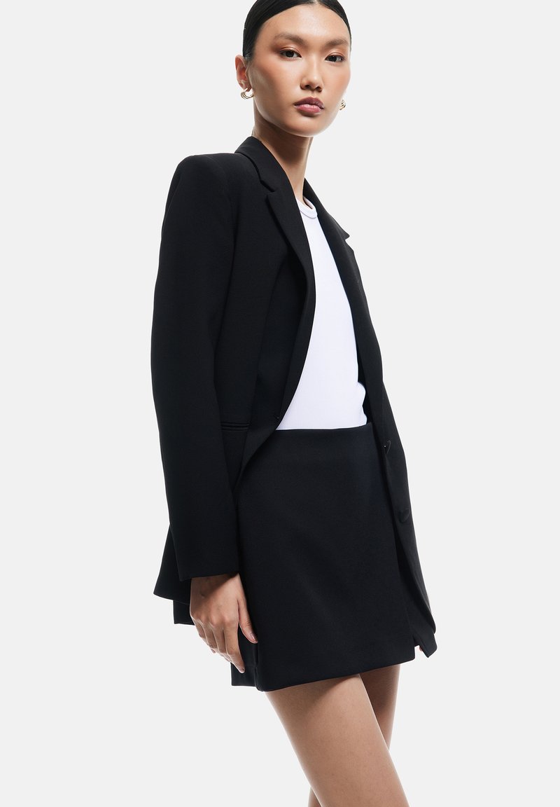 Kvinde iført en sort blazer over en hvid top og en sort mininederdel, stående mod en ensfarvet lys baggrund.