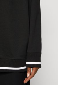 Sudadera negra con puños acanalados que presentan rayas blancas. Textura suave y ajuste relajado con detalle de costura visible en el dobladillo.