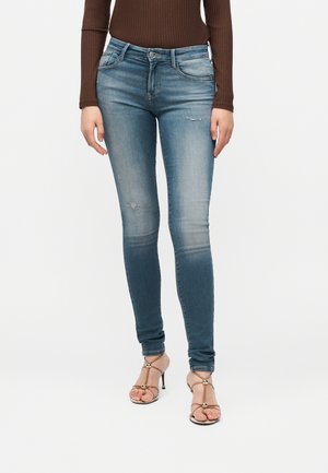 Kvinde iført falmede blå skinny jeans med let slid, brun ribbet langærmet top og beige højhælede sandaler med gyldne accenter.