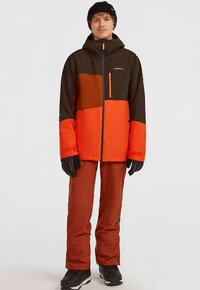 Nuori henkilö, joka on pukeutunut ruskeaan, oranssiin ja punaiseen talvitakkiin, ruosteenvärisiin lumihousuihin, mustiin hanskoihin, mustiin saappaisiin ja mustaan myssyyn.