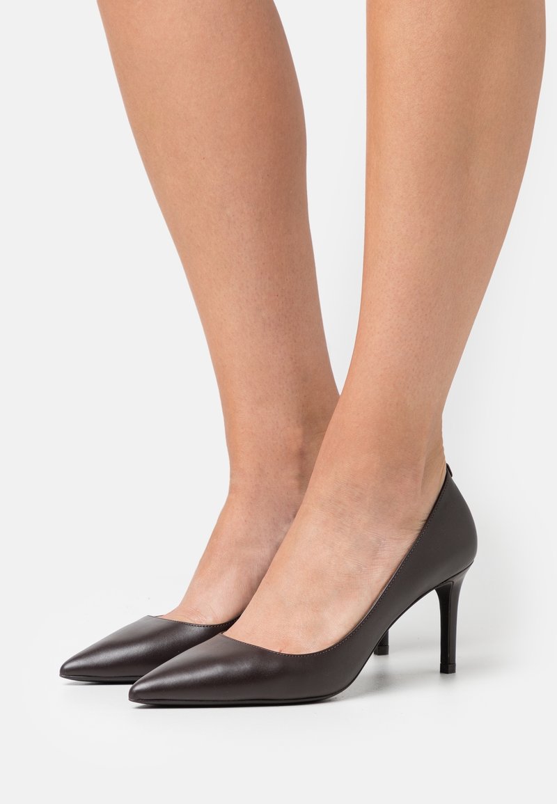 Jambes portant des escarpins en cuir noirs à bout pointu avec des talons aiguille fins sur un fond blanc.