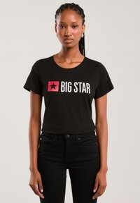 Tânără cu părul împletit, purtând un tricou negru "BIG STAR" și pantaloni negri, pe un fundal simplu și luminos.
